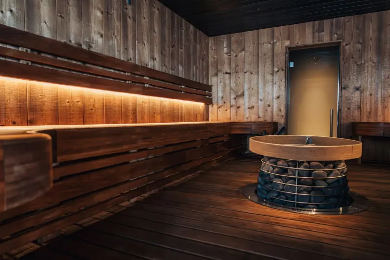 Sauna en bois sombre aux banquettes rétroéclairées d'une lumière chaude, avec un poêle à pierres au centre