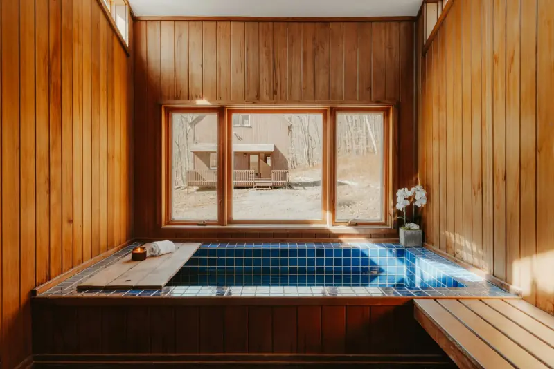 Pièce lambrissée de bois clair avec un bassin carrelé de bleu, un banc en bois et de grandes fenêtres donnant sur la forêt